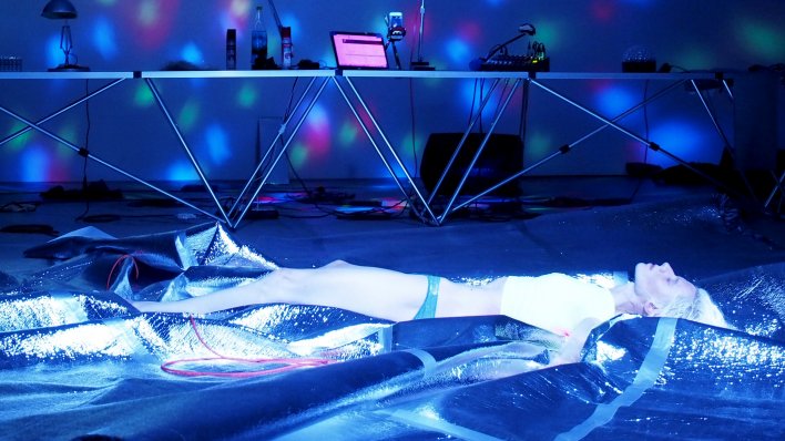 Ein Mensch liegt mit geschlossenen Augen auf einer blau glitzernden Bühne. Im Hintergrund steht ein DJ Pult.