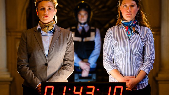 Zwei Frauen vor einer digitalen Zeitanzeige, im Hintergrund ein Mann