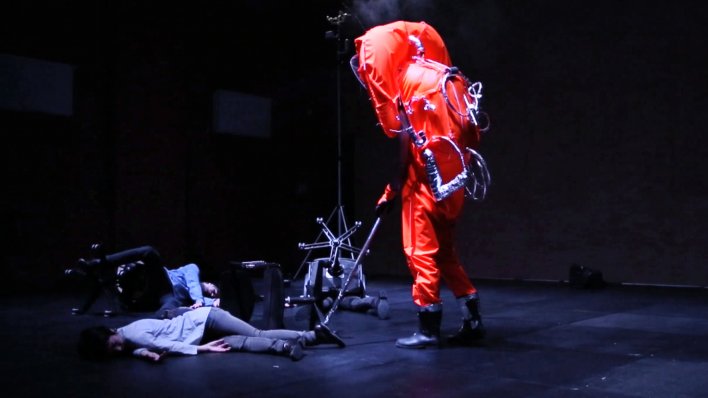 A person is standing on stage in  a space-suit and is cleaning the office.