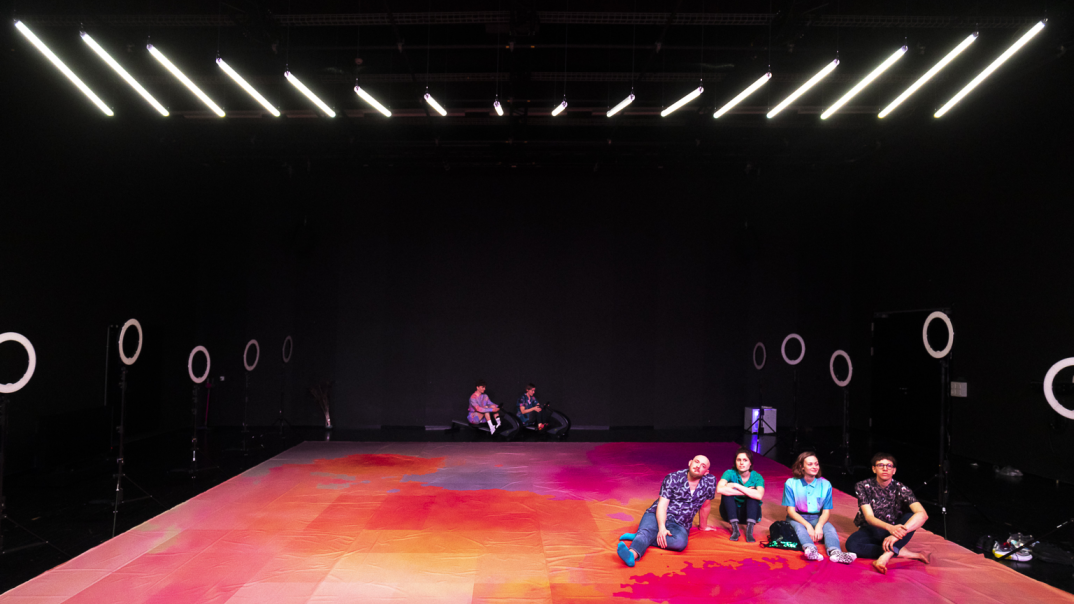 Dieses Foto zeigt die Schauspieler im Aktion. In der Vordergrund sitzen vier Personen lässig und bequem auf dem Boden, während zwei Personen in der Hintergrund ebenfalls sitzen. Das Boden ist mit der Farbe in rote, rosa, orange Töne verseht. 