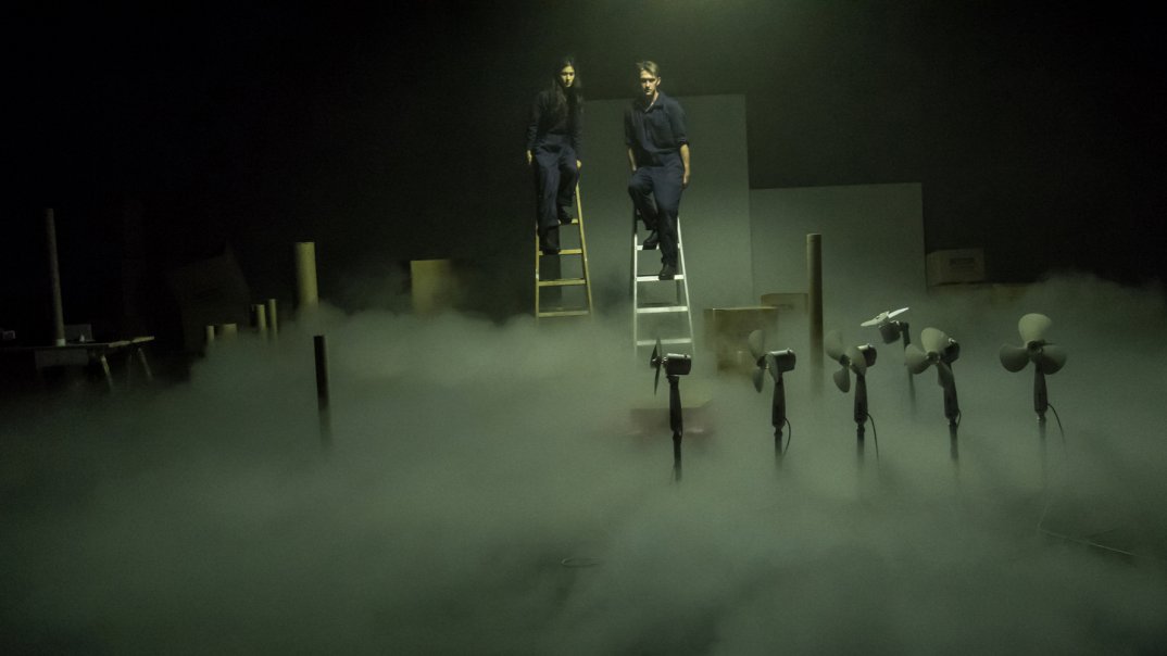 Zwei Personen in dunkelblauen Arbeitskleidung, eine Frau und ein Mann, sind an der hohen Leiter angelehnt und starrten in den dunklen Nebel am Boden. In dem Nebel sind ein paar Ventilatoren in der Reihe aufgestellt.