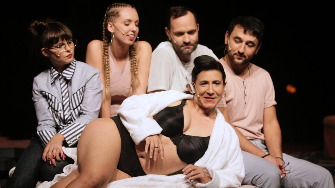 Fünf Personen, drei Frauen und zwei Männer, sitzen auf einen beigen Sofahocker. Eine Frau im Vordergrund ist nur in einen weissen Bademantel und eine schwarze Unterkleidung.